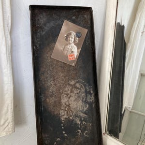 Könnte beinhalten: Ein vintage Backblech aus Metall mit einer abgenutzten, dunkelbraunen Patina. Eine Schwarzweiß-Postkarte eines jungen Mädchens ist mit einem Magneten an das Blech befestigt.