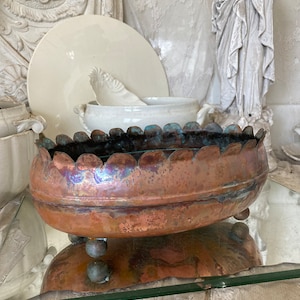 May include: A large, oval copper planter with a scalloped rim and three round feet. The planter has a rustic, aged patina and is sitting on a glass surface.