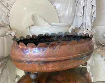 May include: A large, oval copper planter with a scalloped rim and three round feet. The planter has a rustic, aged patina and is sitting on a glass surface.
