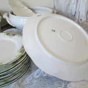 May include: A set of white porcelain plates with a green and purple floral design. The plates are stacked on top of each other. The bottom plate has a scalloped edge.