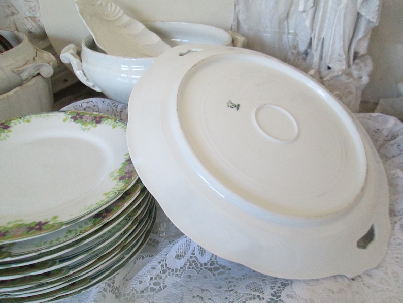 May include: A set of white porcelain plates with a green and purple floral design. The plates are stacked on top of each other. The bottom plate has a scalloped edge.