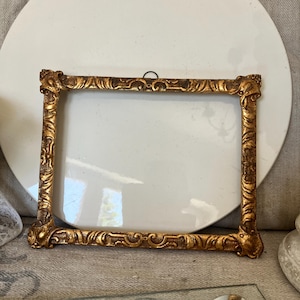 May include: A rectangular picture frame with a metallic gold finish and an ornate, raised relief pattern along all four sides. The frame features decorative corner details and a small black metal hanging loop attached to the top center edge.