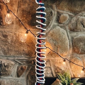 May include: A spiral-shaped hanging decoration crafted from crocheted yarn in alternating stripes of navy blue, red, and white. The long, twisted structure features a wooden bead at the top and a multi-colored yarn tassel at the bottom end.