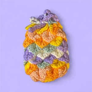 May include: A handmade crochet bag with a dragon scale design. The bag is made from yarn and features a range of colours, including orange, yellow, purple, green, and white. It has a drawstring closure.