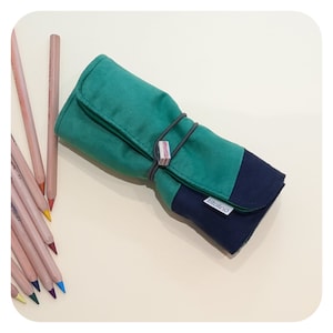 Puede incluir: Un rollo de lápices de color azul marino y verde azulado con una banda elástica y una pequeña etiqueta de madera que dice "titolino".