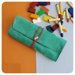 May include: A teal suede pencil case with a red cord closure, holding several crayons. The crayons are in various colours, including yellow, red, and blue. The background is a mix of orange and white.