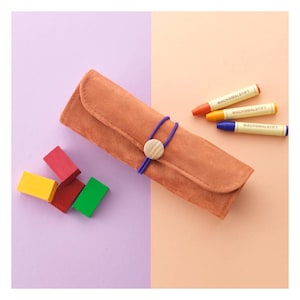 May include: A brown suede pencil case with a purple elastic band and a wooden button. The case is lying on a pink and orange surface. There are four crayons and four wooden blocks in the image. The crayons are labelled "Wachsmalstift".