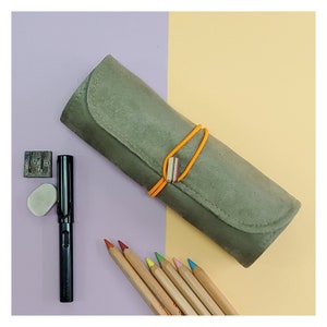 May include: A green suede pencil case with a rubber band closure. The case is rolled up and has a light brown background. There are also a black pen, a white eraser, and a set of colored pencils on the background.