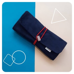 Op de afbeelding: Een donkerblauwe opgerolde potlood etui met een rood elastiek en een zilveren metalen sluiting. De etui heeft het woord "Titolo" erop gedrukt.