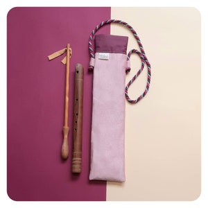 Puede incluir: Una flauta de madera de color marrón y beige, con una funda de transporte de tela rosa con una correa trenzada negra y roja. La funda tiene la palabra "titolano" impresa.