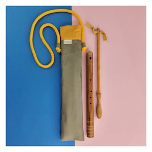 Puede incluir: Una funda de transporte de tela gris con una solapa amarilla y la palabra "titolino" impresa en ella. Una flauta de madera con seis agujeros y un palo de madera con un extremo redondeado están sobre una superficie rosa.