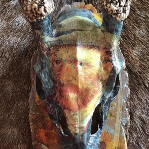 Könnte beinhalten: Ein bemalter Hirschschädel mit einem Porträt von Vincent van Gogh. Das Kunstwerk verwendet eine Palette aus Gelb, Blau und Orange, mit den Geweihen noch befestigt. Der Hintergrund ist eine strukturierte, braune Oberfläche.