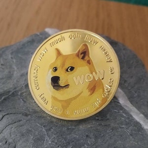 May include: Gold-coloured Dogecoin cryptocurrency coin featuring a Shiba Inu dog with the text "wow much coin how very currency" and "plz mine 4 me" on the left side and "so very good" and "wow" on the right side.