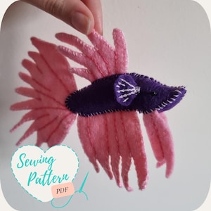 May include: A pink and purple felt sewing pattern for a betta fish. The pattern includes the text "Sewing Pattern PDF".