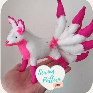 May include: A white and pink felt fox plushie with a nine-tailed fox design. The fox has pink accents on its ears, legs, and tail. The tail is made of nine fluffy white tails with pink tips. The fox is holding a pink flower in its mouth. The plushie is sitting on a hand. The image includes the text "Sewing Pattern PDF".