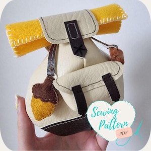 May include: A white felt backpack with brown straps and a yellow felt sleeping bag on top. The backpack has a brown felt acorn attached to the side. The text "Sewing Pattern PDF" is on a pink heart.