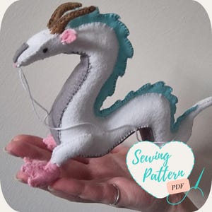 Puede incluir: Un dragón de fieltro blanco con detalles azules y grises. El dragón tiene patas y cuernos rosas. El dragón está siendo sostenido en una mano. La imagen incluye el texto "Sewing Pattern PDF".