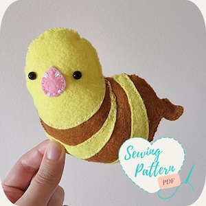 May include: A handmade felt caterpillar toy in yellow and brown, with a pink nose and black eyes. The caterpillar has yellow and brown stripes. The image includes the text "Sewing Pattern PDF" in a heart-shaped graphic.