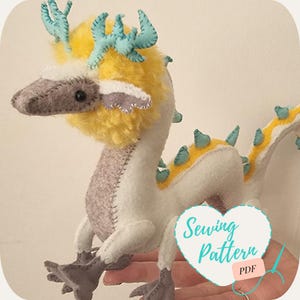 May include: A white and gray felt dragon with yellow fur and turquoise antlers. The dragon has a long tail with turquoise spikes. The dragon is being held in a hand. The image includes the text "Sewing Pattern PDF".