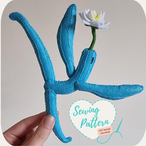 May include: A sewing pattern for a blue felt plush toy shaped like a plant with a white flower on top. The pattern is available as a PDF instant download. The text "Sewing Pattern" is on a pink heart with a needle and thread.
