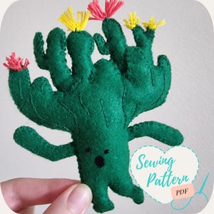 May include: A handmade green felt cactus with yellow and pink flower accents. The cactus has a smiling face and is holding a heart-shaped graphic with the words "Sewing Pattern" and "PDF".