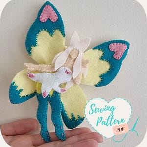 Könnte beinhalten: Ein Nähmuster aus Filz für eine Schmetterlingsfeenpuppe mit einem weißen Kleid, türkisfarbenen Flügeln und rosa Akzenten. Das Muster enthält Anweisungen zum Nähen der Puppe und ist als PDF-Download erhältlich. Nähmuster PDF
