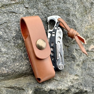 May include: A brown leather multi-tool sheath with a brass snap closure next to a silver and black multi-tool. The multi-tool has a leather lanyard attached. The items are on a gray rock surface.