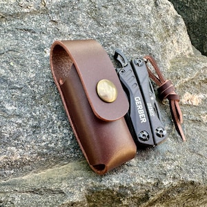 Könnte beinhalten: Ein braunes Lederetui mit Druckknopfverschluss und einem schwarzen Multitool mit einem silbernen Knopf. Das Multitool trägt die Gravur "GERBER".