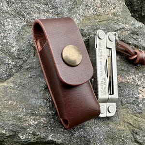 Könnte beinhalten: Eine braune Leder-Multitool-Scheide mit Messingknopfverschluss. Ein silbernes Multitool ist teilweise neben der Scheide zu sehen. Auf dem Multitool steht "LEATHERMAN micra". Eine Lederschnur ist befestigt.