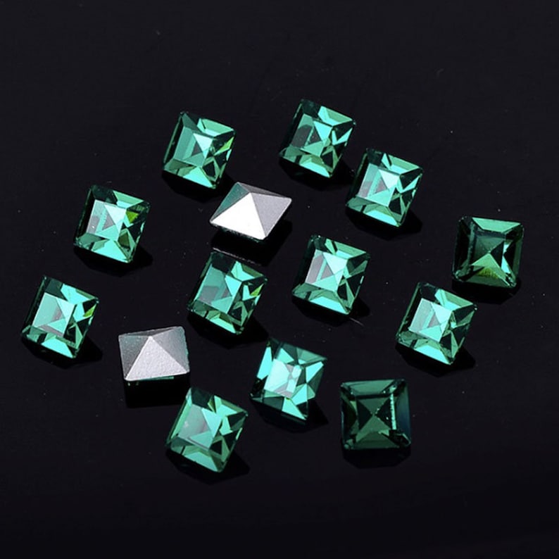May include: A scattering of emerald green square rhinestones on a black background. The rhinestones have a faceted design and are scattered randomly.