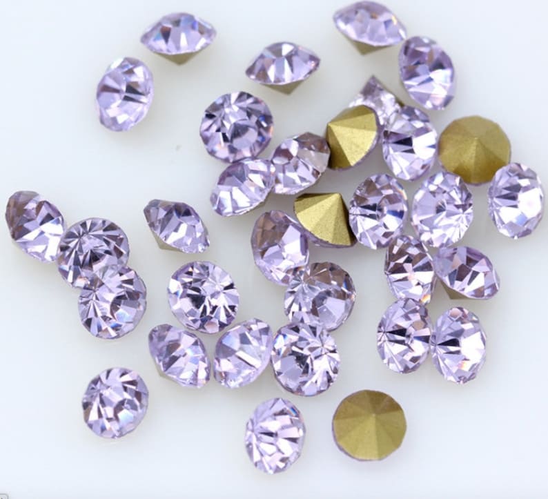 May include: A collection of small, round, lavender-colored rhinestones with a pointed back. The rhinestones are scattered on a white surface.