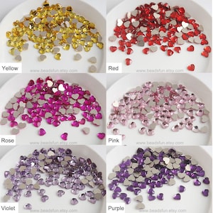 Könnte beinhalten: Sortiment an herzförmigen Strasssteinen in sechs Farben: Gelb, Rot, Rosa, Pink, Violett und Lila. Jede Farbe wird in einer separaten weißen Schale präsentiert. Die Strasssteine sind klein und haben eine flache Rückseite.