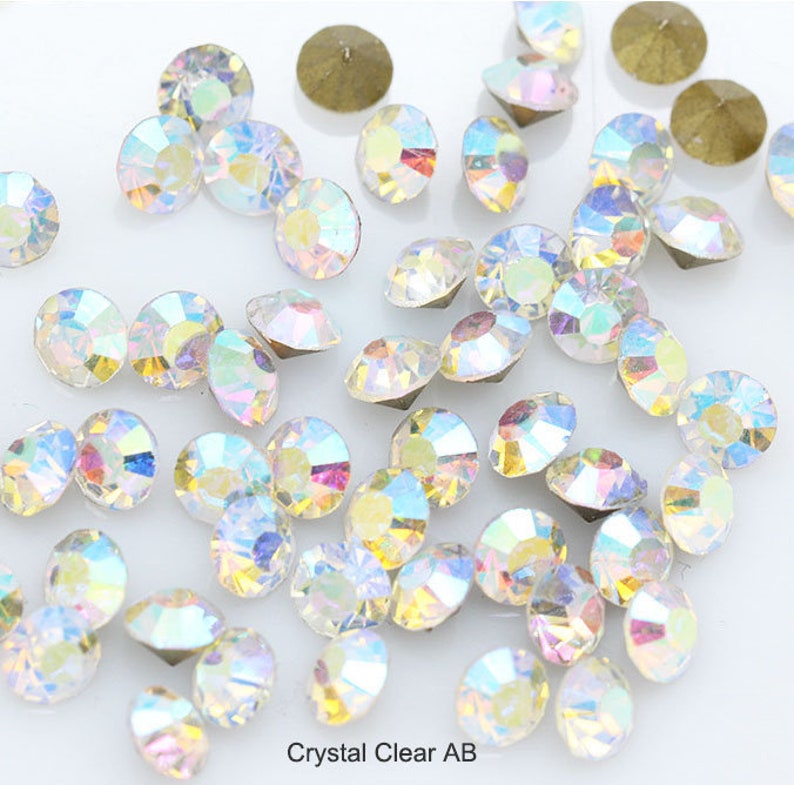Pu&ograve; includere: Una grande quantit&agrave; di strass AB trasparenti iridescenti, sparsi su una superficie bianca. Gli strass sono di varie dimensioni e forme, con un effetto arcobaleno.