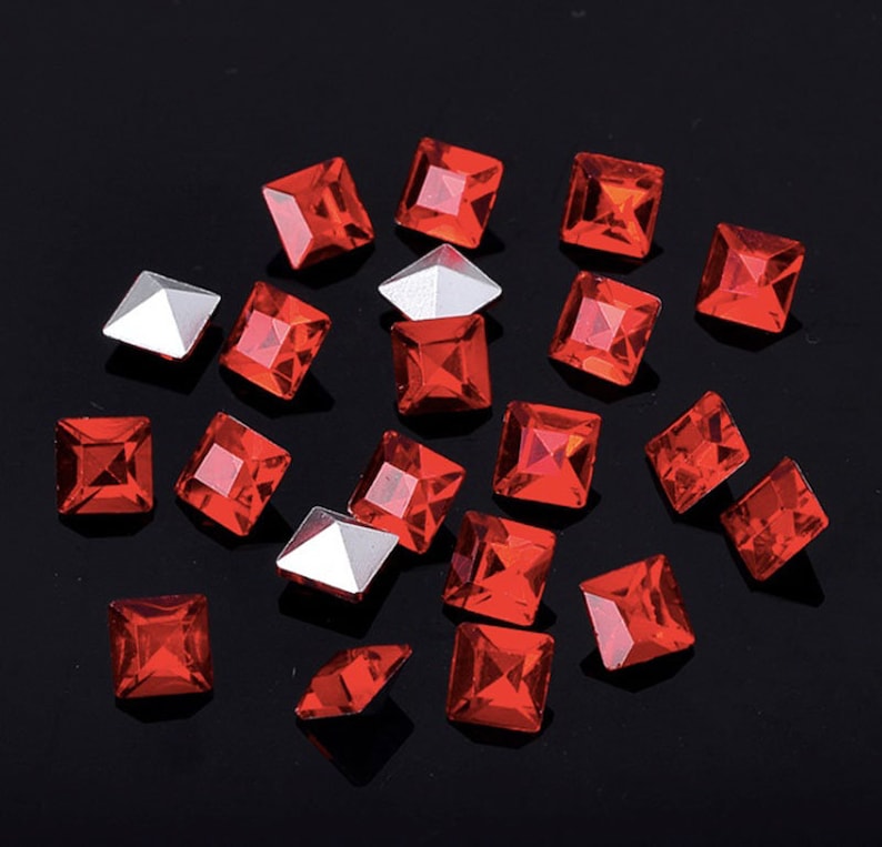 May include: A scattering of red square rhinestones on a black background. The rhinestones are faceted and have a shiny finish.