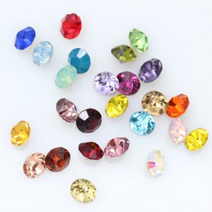 May include: Assortment of colorful round rhinestones in various shades of blue, green, red, yellow, purple, and pink. The rhinestones are scattered on a white surface.