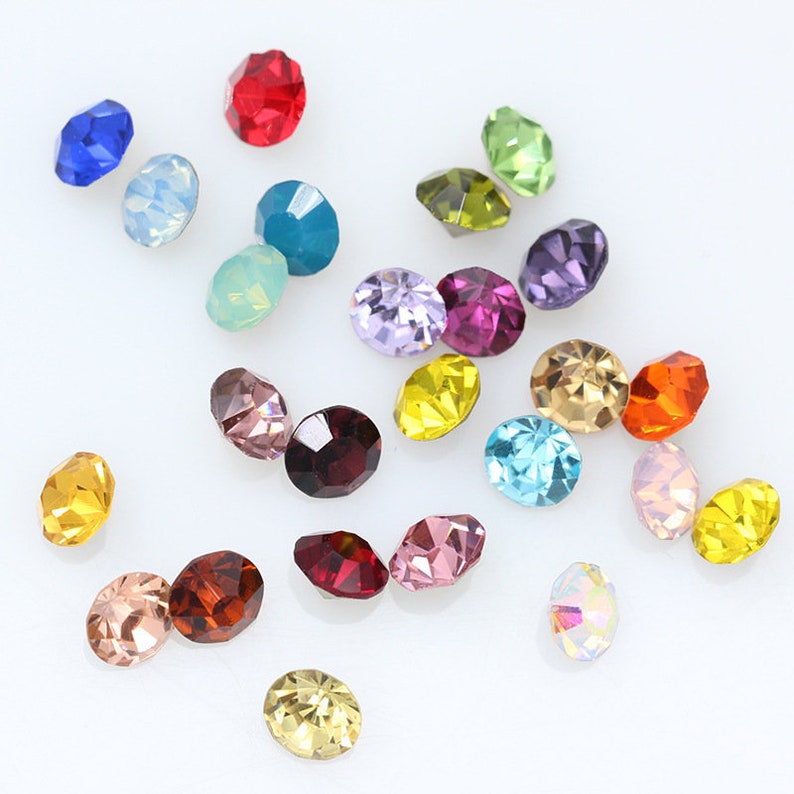 May include: Assortment of colorful round rhinestones in various shades of blue, green, red, yellow, purple, and pink. The rhinestones are scattered on a white surface.