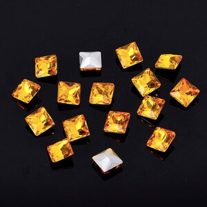 May include: Assortment of small, square, orange-coloured rhinestones on a black background.
