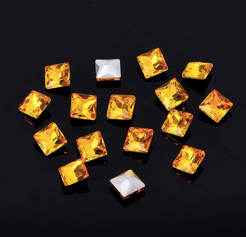May include: Assortment of small, square, orange-coloured rhinestones on a black background.