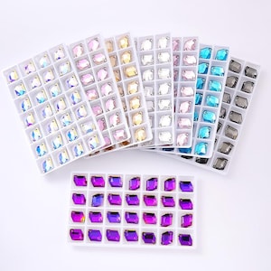 May include: Multiple white plastic trays containing rows of faceted, rectangular-shaped crystals in various colors including iridescent, pink, gold, clear, light pink, blue, dark gray, and vibrant purple, arranged in organized grid compartments.