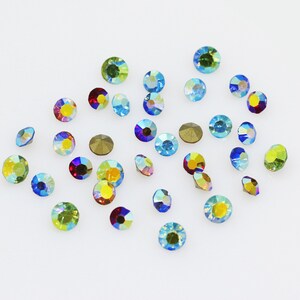 Pu&ograve; includere: Assortimento di strass di vetro iridescenti in vari colori, tra cui blu, verde, rosso e giallo. Gli strass sono sparsi su una superficie bianca.