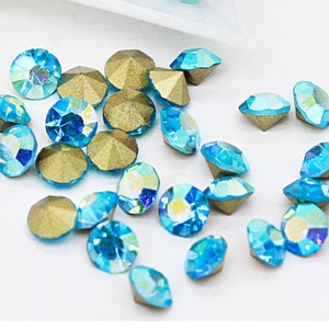 Puede incluir: Surtido de pequeños diamantes de imitación azules iridiscentes redondos con engastes dorados. Los diamantes de imitación tienen un diseño facetado y están dispersos sobre un fondo blanco.