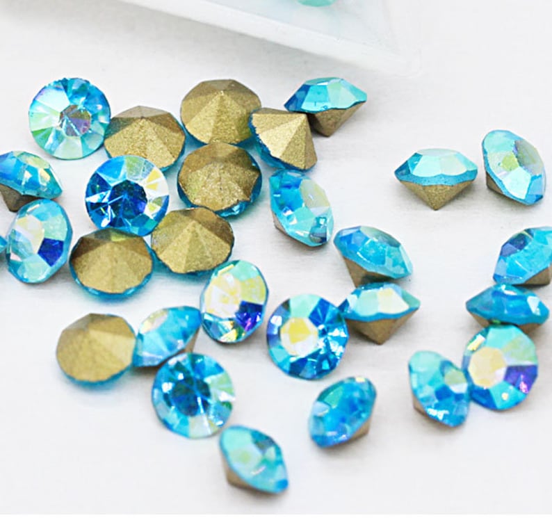 Pu&ograve; includere: Assortimento di piccoli strass rotondi blu iridescenti con montature dorate. Gli strass hanno un design sfaccettato e sono sparsi su uno sfondo bianco.