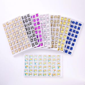 May include: A collection of rectangular plastic trays containing small, square, faceted rhinestones in various colors including gold, silver, purple, pink, yellow, and blue, along with a tray of iridescent, multi-colored square rhinestones.