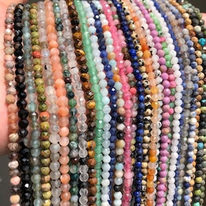 Puede incluir: Una colección de hebras de cuentas en varios colores, incluyendo negro, verde, rosa, azul y marrón. Las cuentas son pequeñas y facetadas, creando un efecto brillante. Las hebras se mantienen juntas, mostrando la variedad de piedras preciosas.