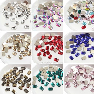 Op de afbeelding: Assortiment van rechthoekige strasssteentjes in verschillende kleuren, waaronder helder, iriserend, rood, blauw, groen, goud en roze. De strasssteentjes worden gepresenteerd op witte borden met een zilveren achterkant.