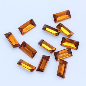 May include: A collection of twelve amber-colored rectangular glass rhinestones. The rhinestones are scattered on a white surface.