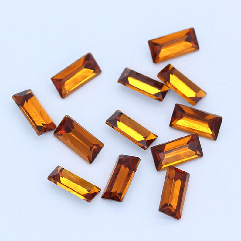 May include: A collection of twelve amber-colored rectangular glass rhinestones. The rhinestones are scattered on a white surface.