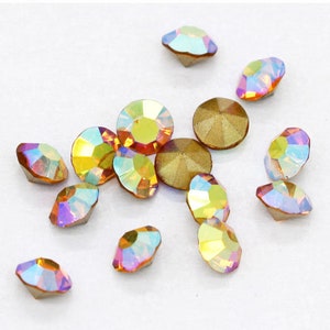 Pu&ograve; includere: Assortimento di strass di vetro iridescenti in varie tonalit&agrave; di oro, arancio e rosa. Gli strass sono sparsi su una superficie bianca.