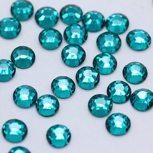 Puede incluir: Cristales de imitación de color azul turquesa, también conocidos como gemas de vidrio, esparcidos sobre una superficie blanca. Estos pequeños cristales redondos y facetados se utilizan para manualidades y decoración.