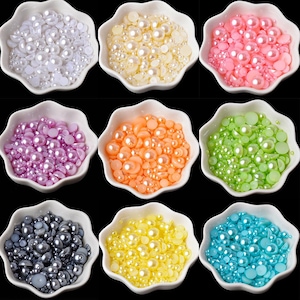May include: A collection of nine white, scalloped-edge bowls, each filled with a different color of flat-backed, half-round faux pearls. The colors include white, cream, pink, purple, orange, lime green, dark gray, yellow, and bright blue.